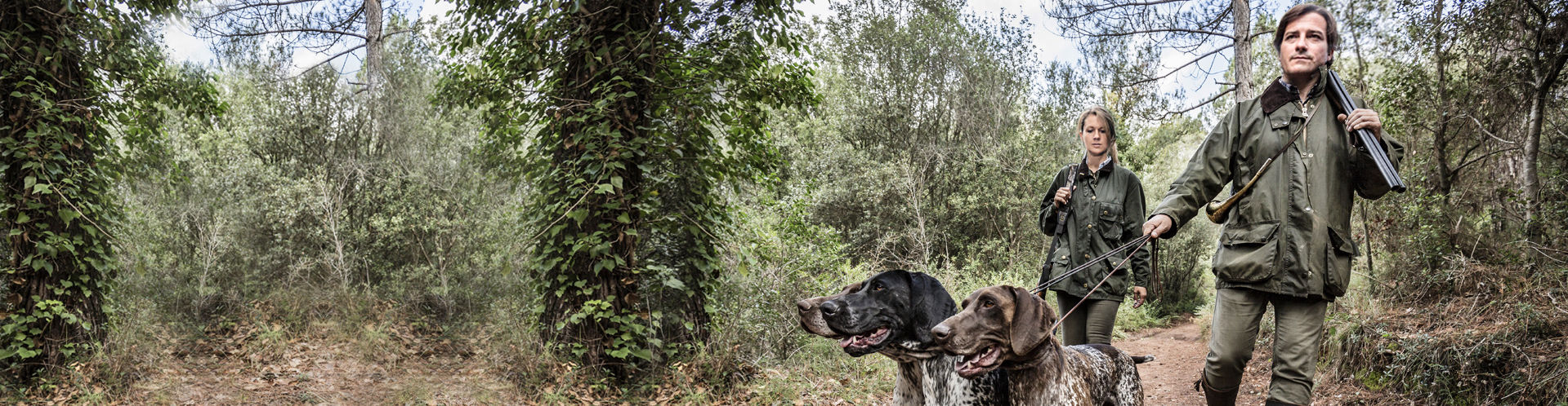 Eine Jägerin und ein Jäger gehen mit ihrer Hundemeute einen Waldweg entlang