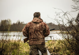 Ein Mann vor einem See in Rückenansicht. Er hält in seinen Händen ein Gewehr