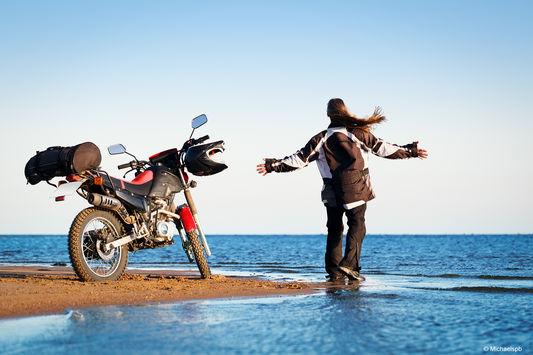 Person breitet ihre Arme vor dem Meer stehend aus und dahinter steht ihr Motorrad.