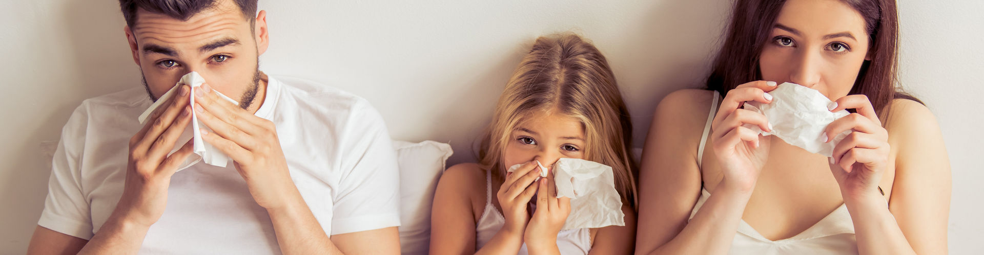 Eine Familie sitzt nebeneinander im Bett und putzt sich die Nase.